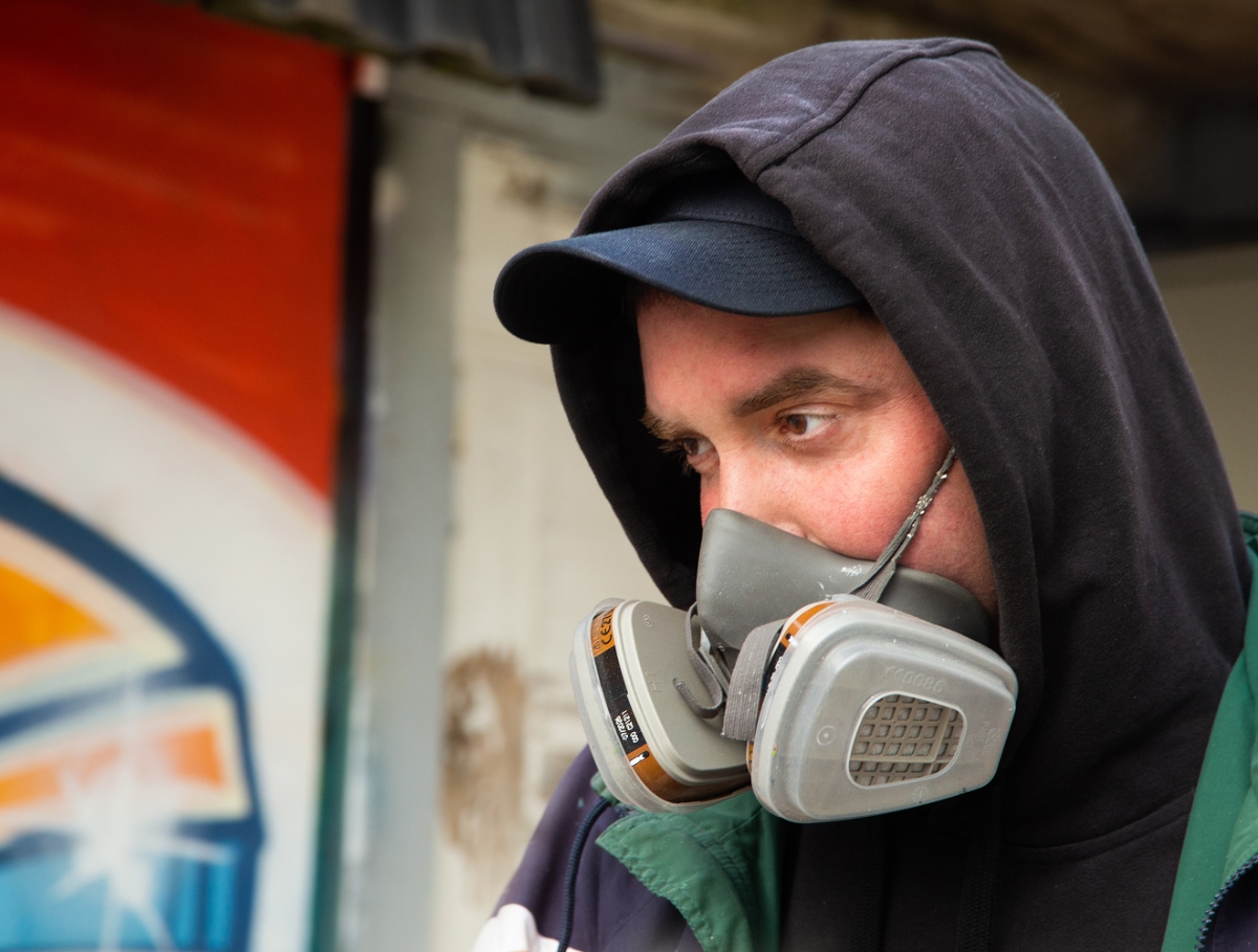 Jon Gredmark with a respirator mask, standing against a wall of graffiti